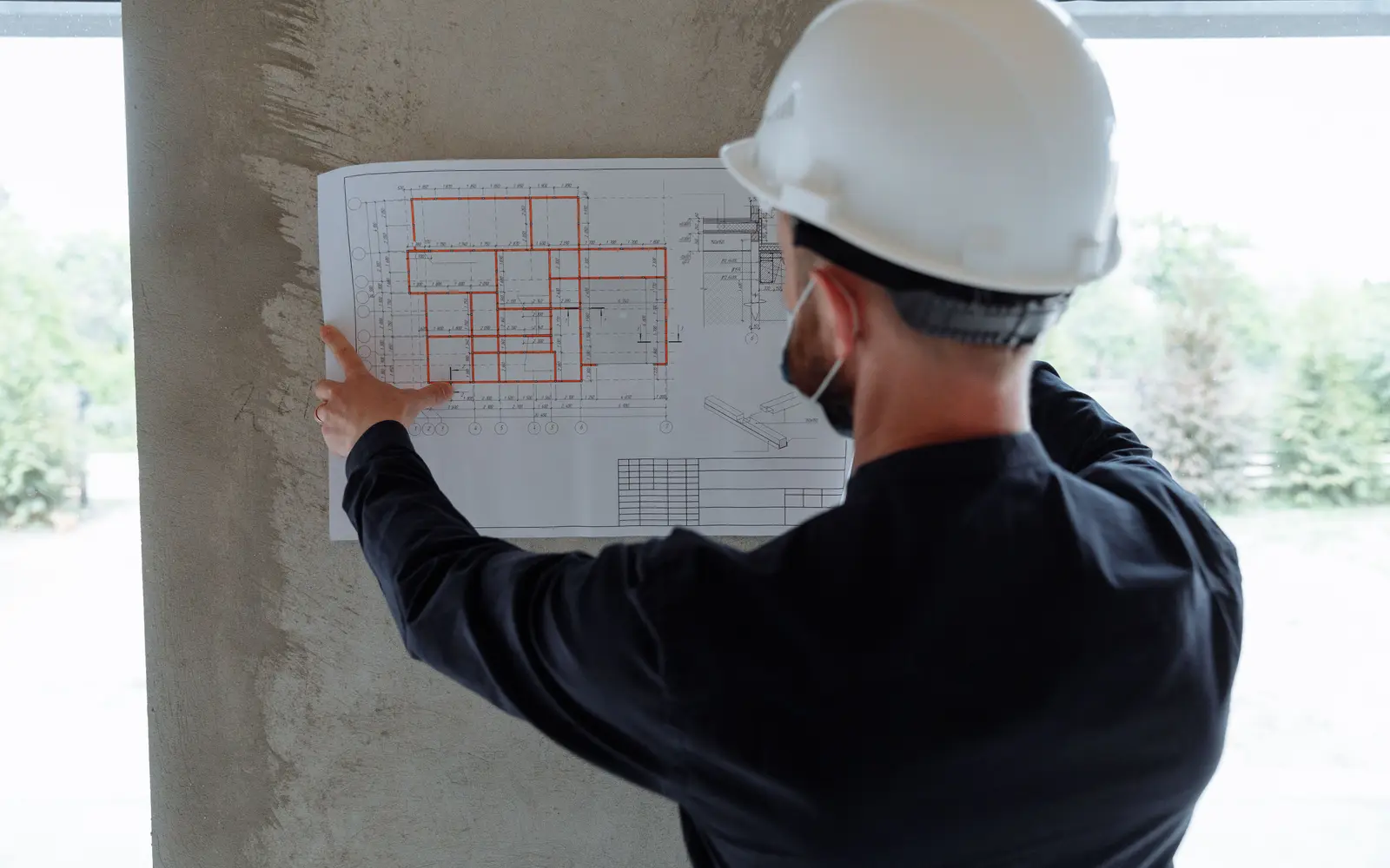 A site engineer in a white hard hat, seen from behind, holding floor plans against a concrete column